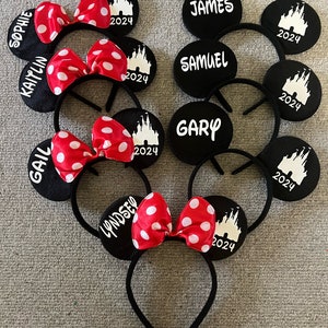 May include: Set of six black mouse ears with red and white polka dot bows. Each ear has a white silhouette of Cinderella's castle and the year 2024. The ears are personalized with names including Sophie, Kaitlyn, Gail, Lyndsey, James, Samuel, and Gary.