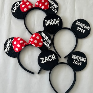May include: Four black mouse ear headbands with red and white polka dot bows. The headbands are labeled "Mommy", "Daddy", "Lucy", and "Zach" with the text "Disneyland 2024" on each headband.