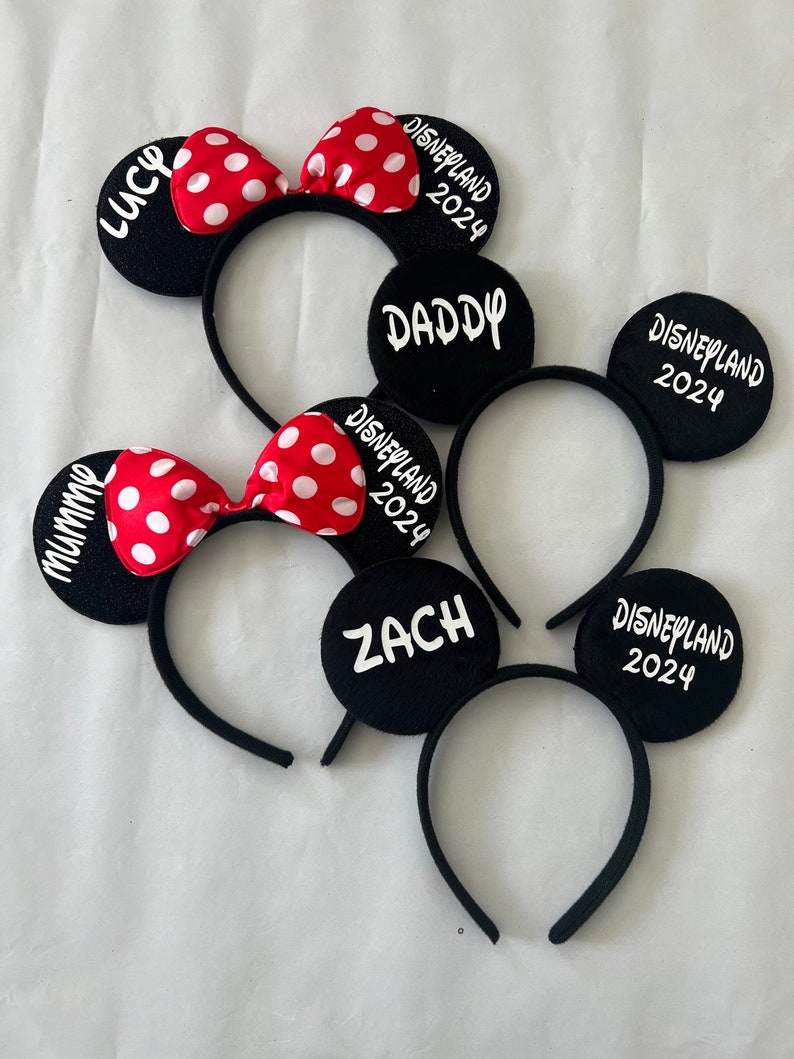 May include: Four black mouse ear headbands with red and white polka dot bows. The headbands are labeled "Mommy", "Daddy", "Lucy", and "Zach" with the text "Disneyland 2024" on each headband.