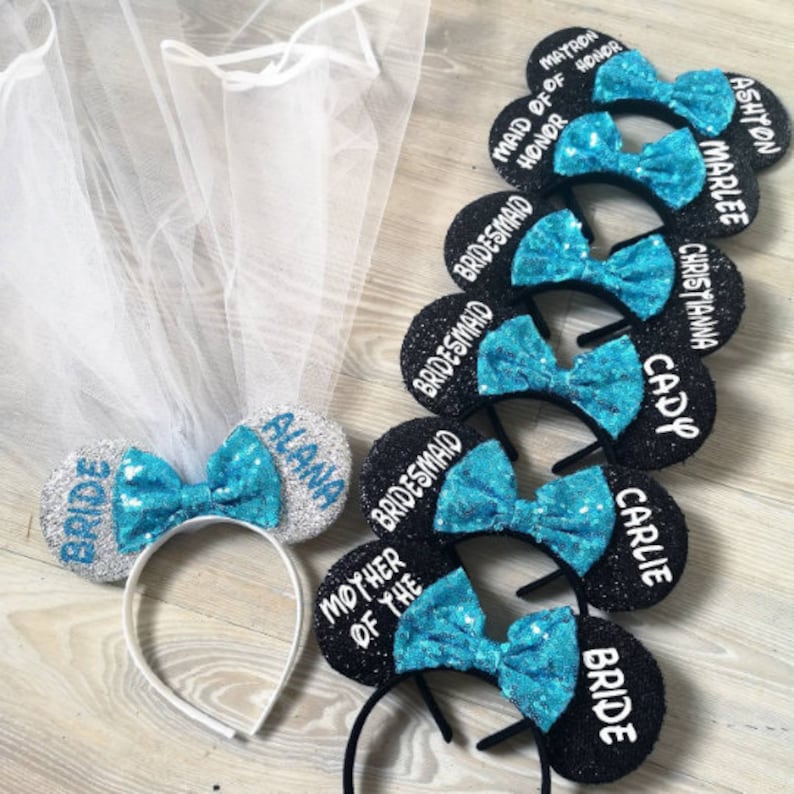 Op de afbeelding: Set van negen zwarte en zilveren muizenoren met blauwe pailletten strikken. Elk oor heeft een andere titel, waaronder "Bride", "Maid of Honor", "Matron of Honor", "Bridesmaid", "Mother of the Bride", "Cady", "Carly", "Christiana" en "Ashyon Marlee".