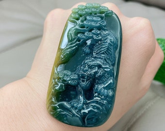 Op de afbeelding: Een groene en gele jade hanger gegraveerd met een tijger en een boom. De hanger is een traditioneel Chinees ontwerp.