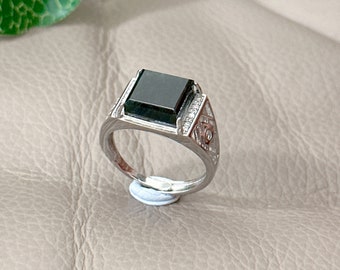 Op de afbeelding: Een zilveren ring met een zwarte vierkante edelsteen in het midden. De ring heeft een gedetailleerd ontwerp met kleine diamanten rond de edelsteen.