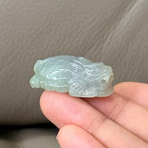 May include: A jade green turtle figurine carved from jade. The turtle is lying on its belly with its head raised. The turtle has detailed carvings on its shell and head.