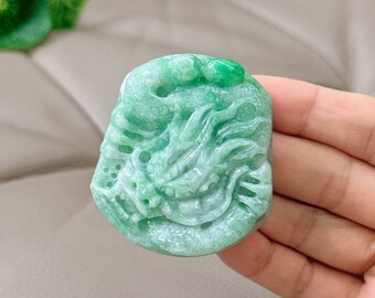 Op de afbeelding: Een groene jade hanger gegraveerd met een gedetailleerd ontwerp van een draak. De hanger heeft een glad, gepolijst oppervlak en is een prachtig voorbeeld van jade-snijwerk.