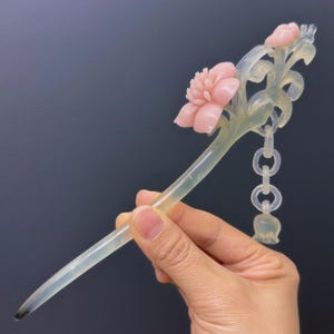 Puede incluir: Horquilla decorativa con un tallo verde claro y una flor rosa. El accesorio para el cabello tiene una cadena de pequeños eslabones transparentes y un pequeño dije transparente en forma de campana. La horquilla se sostiene en una mano.