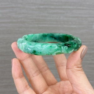 May include: A green jade bangle bracelet with intricate carvings. The bracelet is carved with a floral design and is a beautiful example of traditional Chinese craftsmanship.