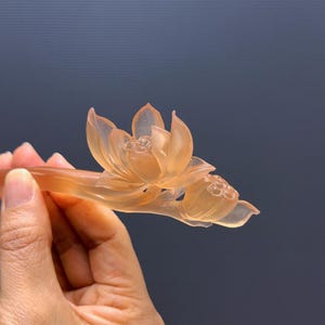 Puede incluir: Un palillo para el pelo con forma de flor de loto naranja transl&uacute;cido. El accesorio para el pelo presenta una talla detallada de una flor de loto en flor y un capullo cerrado. El palillo para el pelo se sostiene sobre un fondo azul oscuro.