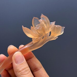 Puede incluir: Una horquilla de color melocot&oacute;n transl&uacute;cido tallada en forma de flor de loto. La flor tiene p&eacute;talos detallados y un capullo, con un tallo largo y delgado. El accesorio para el cabello se sostiene sobre un fondo oscuro.