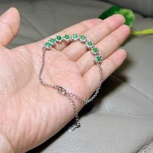 Op de afbeelding: Een zilveren armband met een rij ovale groene edelstenen, omringd door kleinere heldere edelstenen. De armband heeft een karabijn sluiting.