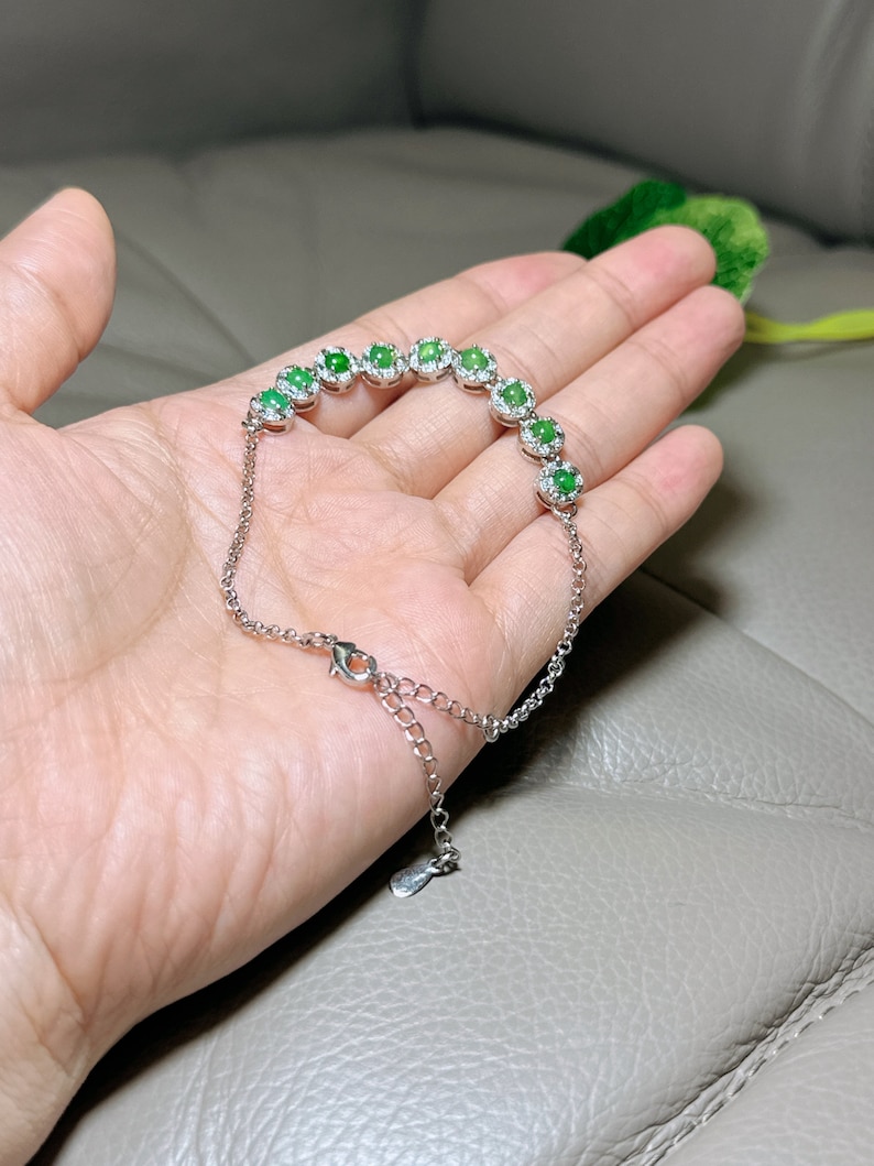 Op de afbeelding: Een zilveren armband met een rij ovale groene edelstenen, omringd door kleinere heldere edelstenen. De armband heeft een karabijn sluiting.