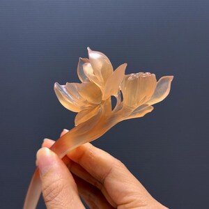 Puede incluir: Una horquilla de color melocot&oacute;n transl&uacute;cido con un dise&ntilde;o detallado de flor de loto. La flor tiene m&uacute;ltiples p&eacute;talos y un tallo delicado, creando un accesorio elegante. La horquilla se sostiene sobre un fondo oscuro.