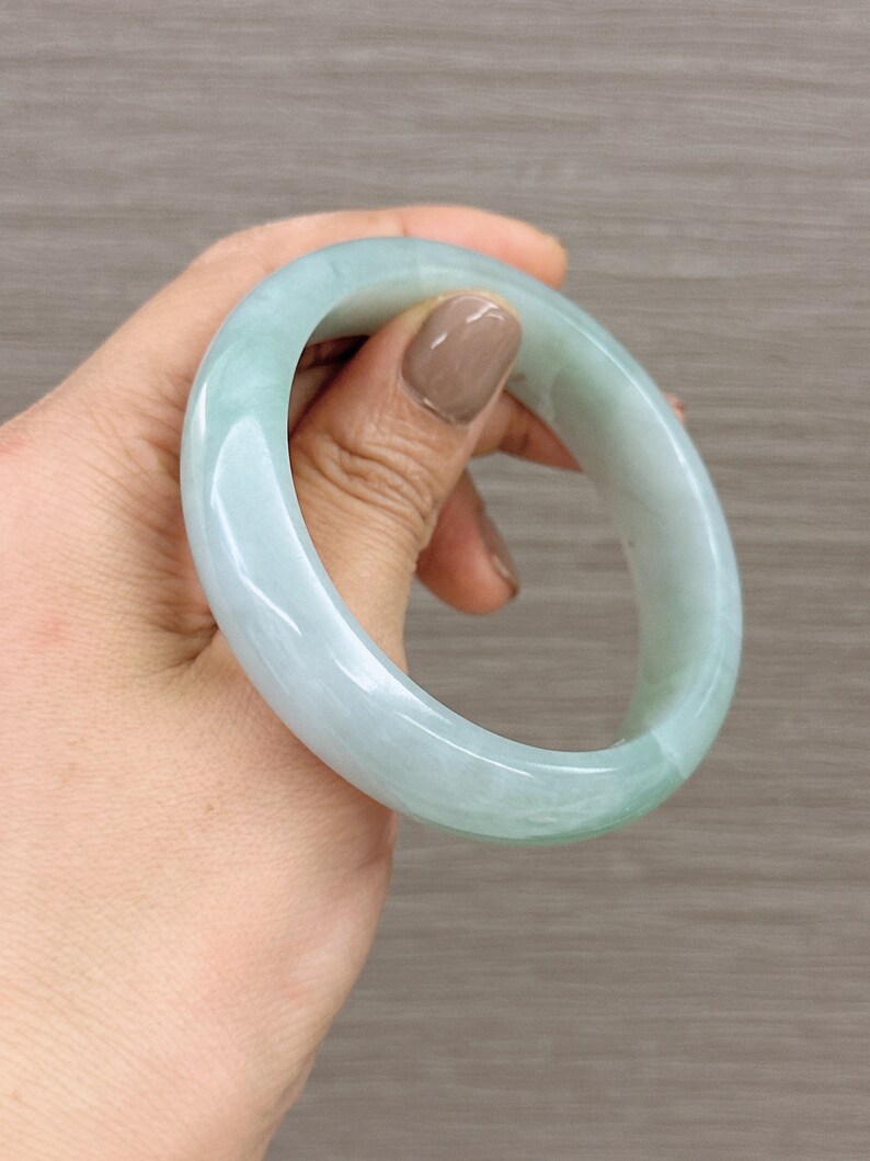 May include: A light green jade bangle bracelet. The bangle is smooth and polished, with a subtle swirl pattern.