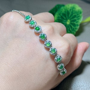 Op de afbeelding: Een zilveren armband met een kettingontwerp heeft een rij ronde groene edelstenen omringd door kleinere heldere edelstenen.
