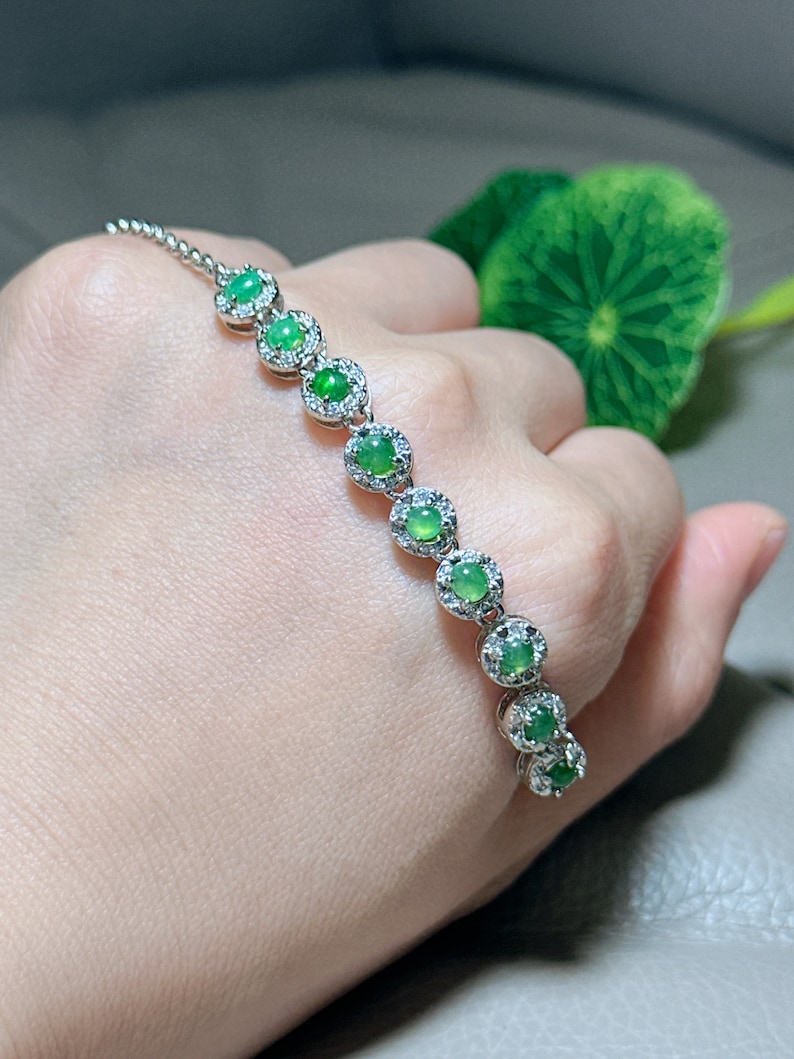 Op de afbeelding: Een zilveren armband met een kettingontwerp heeft een rij ronde groene edelstenen omringd door kleinere heldere edelstenen.