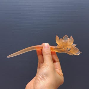 Puede incluir: Un palillo para el pelo con forma de flor de loto naranja transl&uacute;cida. El accesorio para el cabello presenta un tallo curvo y un dise&ntilde;o detallado de flor de loto. El palillo para el pelo se sostiene sobre un fondo gris oscuro.