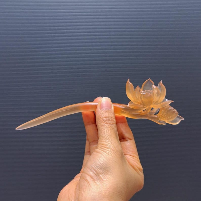 Puede incluir: Un palillo para el pelo con forma de flor de loto naranja transl&uacute;cida. El accesorio para el cabello presenta un tallo curvo y un dise&ntilde;o detallado de flor de loto. El palillo para el pelo se sostiene sobre un fondo gris oscuro.