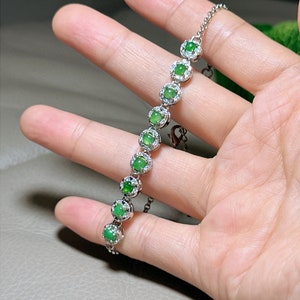 Op de afbeelding: Een zilveren armband met een ketting en groene edelstenen die in zilver zijn gezet. Elke edelsteen is omgeven door kleinere, heldere stenen.