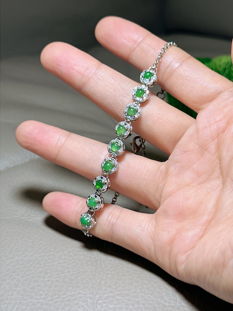 Op de afbeelding: Een zilveren armband met een ketting en groene edelstenen die in zilver zijn gezet. Elke edelsteen is omgeven door kleinere, heldere stenen.