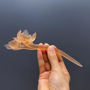 Puede incluir: Horquilla transl&uacute;cida de color &aacute;mbar con un delicado dise&ntilde;o de flor de loto. El accesorio para el cabello se sostiene sobre un fondo gris oscuro, mostrando los intrincados detalles del tallado floral. La horquilla mide aproximadamente 15 cm de largo.