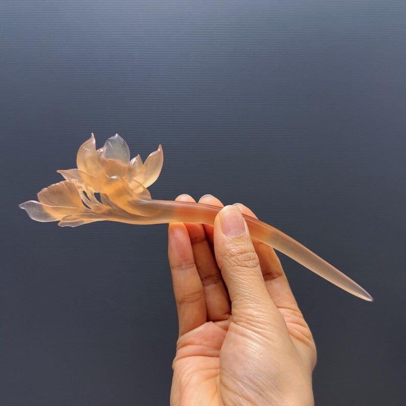 Puede incluir: Horquilla transl&uacute;cida de color &aacute;mbar con un delicado dise&ntilde;o de flor de loto. El accesorio para el cabello se sostiene sobre un fondo gris oscuro, mostrando los intrincados detalles del tallado floral. La horquilla mide aproximadamente 15 cm de largo.
