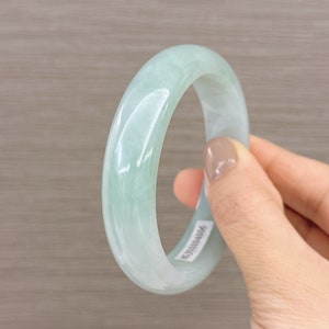 May include: A light green jade bangle bracelet. The bangle is smooth and polished. The bangle has a sticker on it with the number "BJ01034556".