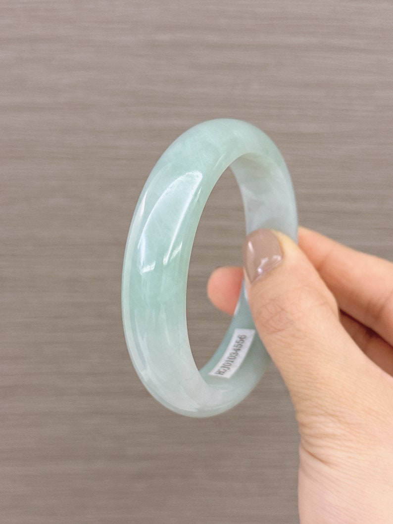 May include: A light green jade bangle bracelet. The bangle is smooth and polished. The bangle has a sticker on it with the number "BJ01034556".