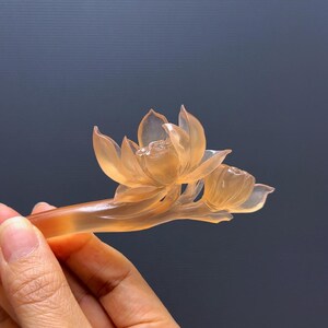 Puede incluir: Un palillo para el pelo con forma de flor de loto naranja transl&uacute;cida. La flor est&aacute; intrincadamente tallada con delicados p&eacute;talos y un capullo. El accesorio para el pelo se sostiene en una mano sobre un fondo oscuro.