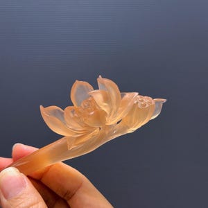 Puede incluir: Una horquilla con forma de flor de loto naranja transl&uacute;cida. La flor est&aacute; intrincadamente tallada con p&eacute;talos detallados y un delicado tallo. El accesorio para el cabello se sostiene sobre un fondo azul oscuro.