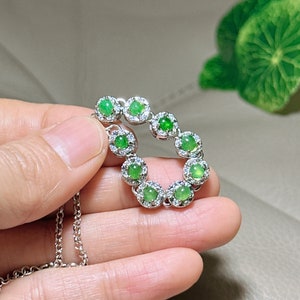 Op de afbeelding: Een zilveren ketting met een hanger met een cirkel van groene jade stenen omringd door kleinere heldere stenen.