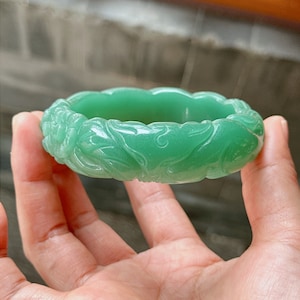 May include: A green jade bangle with intricate floral carvings. The bangle is smooth and polished, with a delicate design.