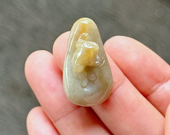 Puede incluir: Un colgante de jade tallado con un diseño detallado de una rana. El colgante es de color verde claro con acentos amarillos. Tiene forma de lágrima y un acabado liso y pulido.
