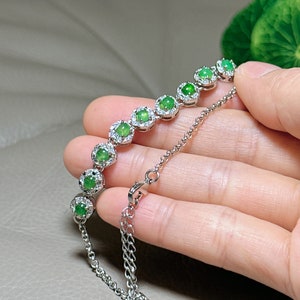 Op de afbeelding: Een zilveren armband met een ketting en groene edelsteenaccenten. Elke edelsteen is omgeven door een halo van kleinere heldere stenen.