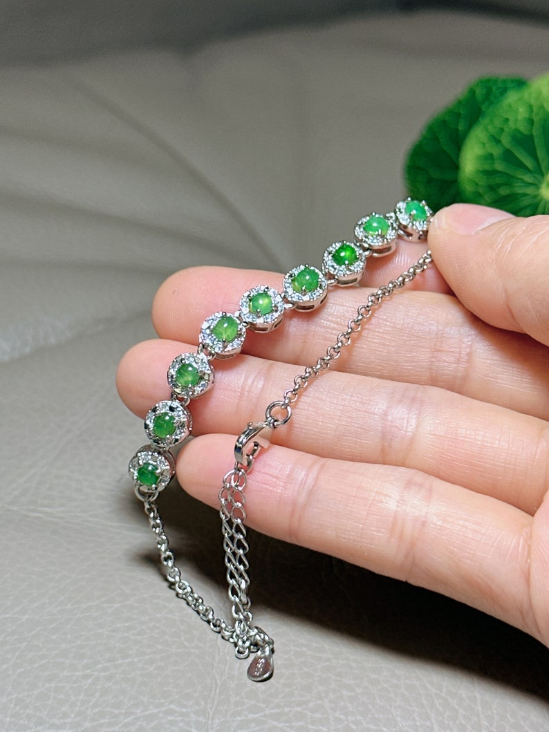 Op de afbeelding: Een zilveren armband met een ketting en groene edelsteenaccenten. Elke edelsteen is omgeven door een halo van kleinere heldere stenen.