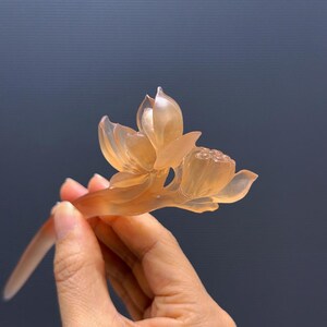 Puede incluir: Un palillo para el pelo con forma de flor de loto transl&uacute;cida de color &aacute;mbar. La flor est&aacute; intrincadamente tallada con delicados p&eacute;talos y una detallada vaina de semillas. El tallo es largo y delgado, dise&ntilde;ado para insertarse en el cabello.