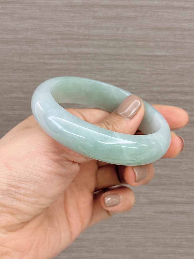 May include: A light green jade bangle bracelet. The bangle is smooth and polished, with a subtle marbled pattern.