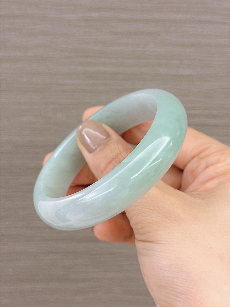 May include: A light green jade bangle bracelet. The bangle is smooth and polished, with a subtle swirl pattern.