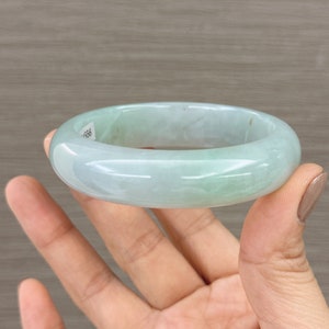 May include: A light green jade bangle bracelet. The bracelet is smooth and polished, with a subtle marbling effect. The bracelet is held in a hand, with the fingers visible.