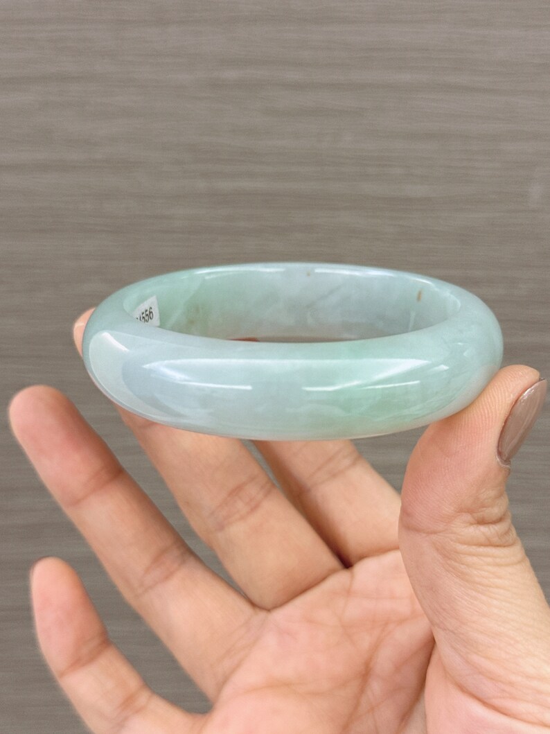 May include: A light green jade bangle bracelet. The bracelet is smooth and polished, with a subtle marbling effect. The bracelet is held in a hand, with the fingers visible.