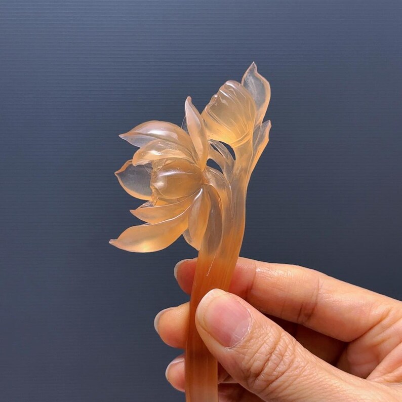 Puede incluir: Un palillo para el pelo transl&uacute;cido, color &aacute;mbar, tallado en forma de flor de loto. Los p&eacute;talos est&aacute;n delicadamente detallados, y el tallo se sostiene en una mano. El fondo es azul oscuro. Accesorio para el cabello decorativo &uacute;nico.