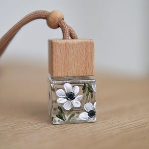 May include: A small glass bottle with a wooden top and a string for hanging. The bottle is decorated with hand-painted white flowers with black centers.