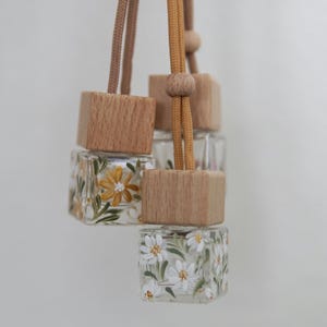 May include: Clear glass air fresheners with wooden tops and floral designs. The air fresheners are square-shaped and suspended by tan and brown cords. One has yellow flowers, and the other has white flowers.