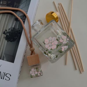 May include: Clear glass diffuser bottle and car air freshener with pink and white floral designs. The diffuser has a gold cap and wooden reeds. The car air freshener has a wooden top and a tan cord.