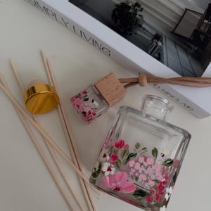 May include: A clear glass diffuser bottle with a floral design in pink and white, alongside wooden reeds and a small matching car air freshener. A gold-coloured cap and a book titled "SIMPLY LIVING" are also visible.