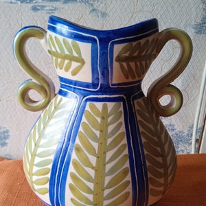 May include: A white ceramic vase with blue and green painted designs. The vase has two handles and a rounded body.