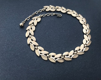 May include: A gold-toned necklace featuring a leaf design. The necklace has a series of connected leaf-shaped elements, creating a continuous pattern. The necklace is adjustable and has a hook closure. The jewelry is displayed on a dark background.