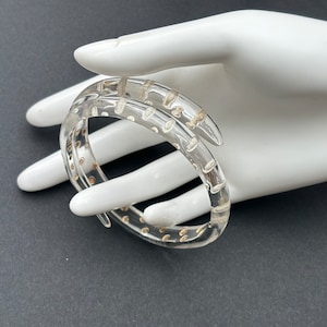 Pode incluir: Pulseira transparente e clara com um design em espiral. A pulseira tem pequenos detalhes circulares dourados. A pulseira é mostrada em uma mão branca sobre um fundo cinza escuro. Uma joia única.