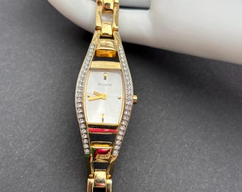 Può includere: Un orologio da polso color oro con quadrante bianco e lunetta tempestata di diamanti. Il quadrante dell'orologio presenta la scritta "Accurist" in lettere nere.