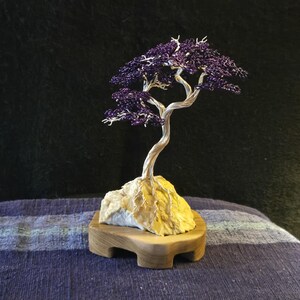 May include: A handcrafted wire tree sculpture with a purple canopy and silver trunk. The tree is mounted on a light-colored rock base, which is set on a wooden platform. The sculpture is a unique decorative item.
