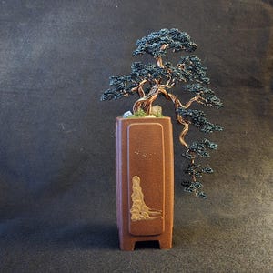 Op de afbeelding: Handgemaakte bonsai boomsculptuur van draad, met donkerblauw blad en een koperkleurige stam. De boom staat in een bruine rechthoekige pot met een decoratief reliëf. Een uniek kunstwerk voor in huis.