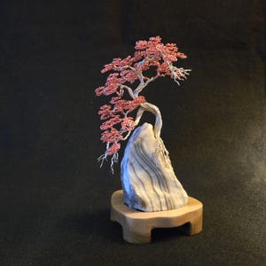 May include: A handcrafted wire bonsai tree sculpture with copper-colored leaves and a silver trunk. The tree is mounted on a gray stone base and a wooden stand, creating a miniature landscape. The sculpture is set against a dark background.